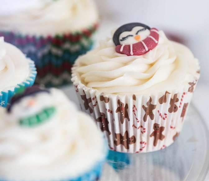 A close-up of a frosted cupcake in a white paper liner decorated with brown gingerbread figures and red candy cane motifs, topped with a penguin figurine wearing a red striped scarf. Other cupcakes are blurred in the background.