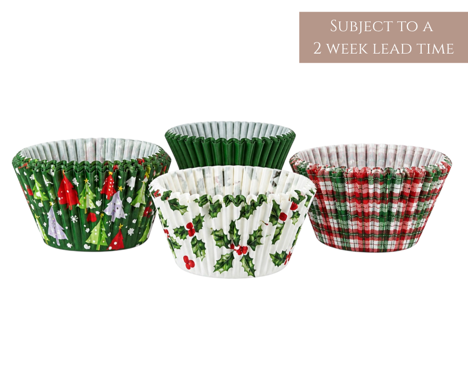 Three festive paper cupcake liners on a white background, featuring a green Christmas tree print, a green holly and berry pattern, and a red and green plaid design. A brown text box in the top right reads 'SUBJECT TO A 2 WEEK LEAD TIME'.
